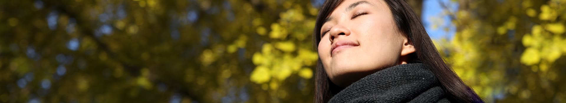 Person outdoors with eyes closed and fall leaves and colors in background.