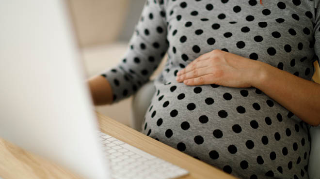 Pregnant woman with hand over belly, sitting and looking at computer screen with keyboard on desk